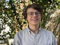 A person standing outdoors, wearing a light-colored button-up shirt. The background features green foliage with sunlight filtering through the leaves.