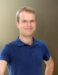 A person standing against a plain background, wearing a blue polo shirt.