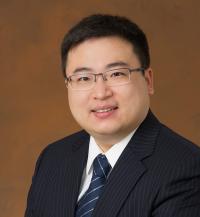 A person wearing a dark pinstripe suit, white dress shirt, and a blue tie with diagonal stripes. The background is a solid brown color. 