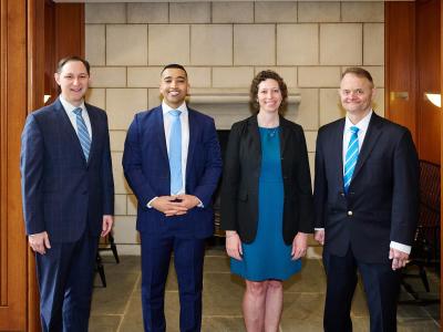 four alumni in suits line up against brick wall