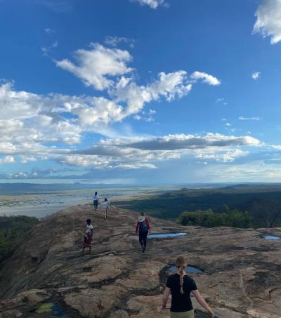 Several people walking on a large rocky surface with shallow puddles, overlooking a vast green landscape and a distant body of water under a bright blue sky with scattered clouds.