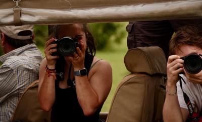 Three people seated in an open safari vehicle, two of them holding cameras and taking photographs, with green vegetation visible in the background.