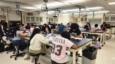 Students seated at lab tables in a science classroom, writing and working with lab materials.