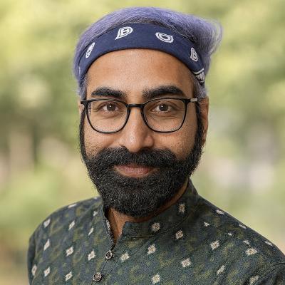 A person wearing a dark green patterned kurta with a high collar and metallic buttons. The individual has purple hair and is wearing a black bandana with white paisley designs. The background is outdoors with greenery and blurred foliage.