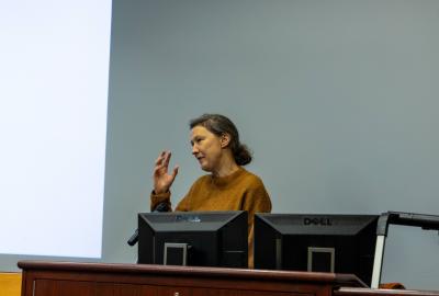 A person standing behind a podium with computer monitors, gesturing during a presentation in a classroom or conference setting.