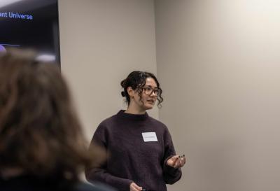Person presenting in a room with a screen showing text, wearing a dark sweater and name tag.