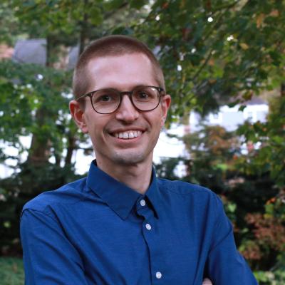 A person in a blue button-up shirt stands outdoors with trees and foliage in the background.