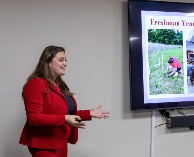 Presenter in a red suit giving a talk beside a screen showing freshman-year fieldwork photos.