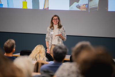 A presenter stands at the front of a lecture hall speaking to an audience, with slides displayed on a large screen behind them.