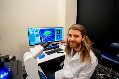 Person in a laboratory seated next to a microscope and computer monitors displaying colorful scientific images.