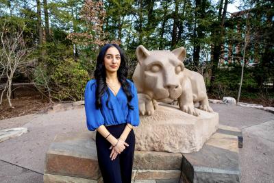 Person posing beside a stone lion sculpture outdoors, surrounded by trees and campus architecture.