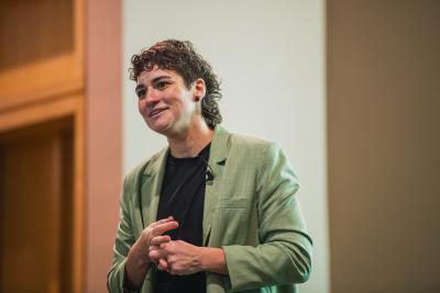 Person in a green blazer speaks on stage, hands clasped, smiling during a professional presentation.