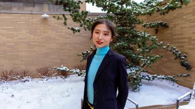 Person standing outdoors in a courtyard with snow on the ground and evergreen trees behind a brick wall.
