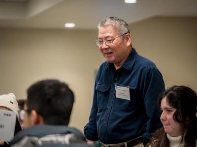 Person wearing glasses and a dark button-up shirt with a name badge stands indoors, leaning slightly forward while speaking to seated people at a table; a classroom or meeting room setting with overhead lighting.