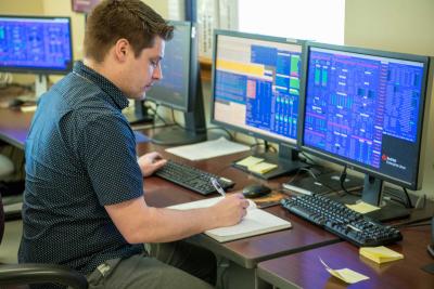 man writes at notebook at a desk with many computer monitors displaying data
