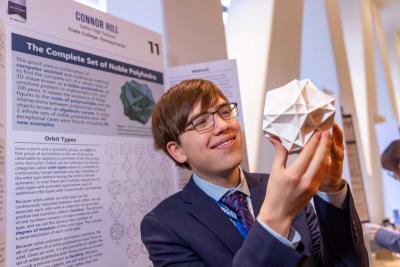 Connor Hill holding model of noble polyhedron in front of his research poster