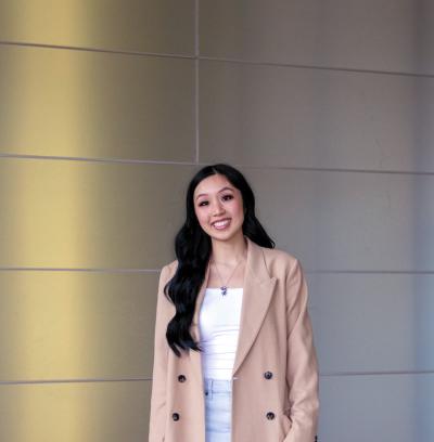 Person in a beige blazer and white top standing against a paneled wall, facing the camera.