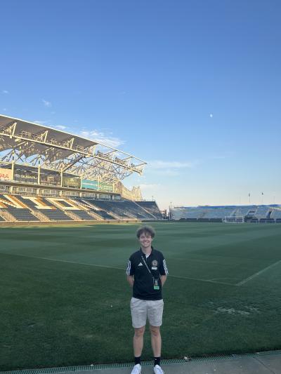 Person standing on a soccer field inside a large stadium under a clear blue sky.