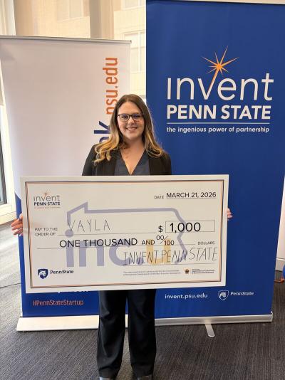 Maya Anderson, Eberly College of Science alumna, posing with a large check fro $1,000 dollars in front of a sign with Invent Penn State's logo.
