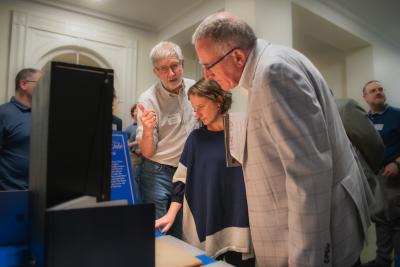 Attendees gather around a display, examining a demonstration and discussing materials at an indoor event.