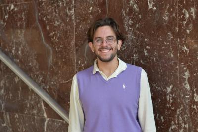 Portrait of a person wearing glasses and a lavender sweater vest, standing in front of a marble wall.