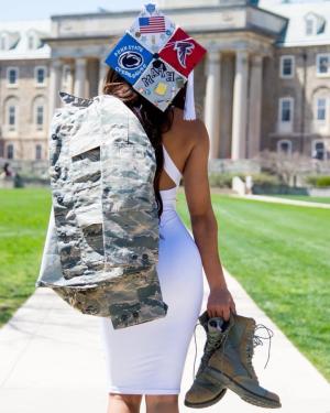 Haley McClain Hill standing while facing Old Main with her graduation cap decorated with Penn State cheerleading, the American flag, the Atlanta Falcons' logo, and math. She's holding her Air Force uniform over her shoulder and her boots in her other hand.