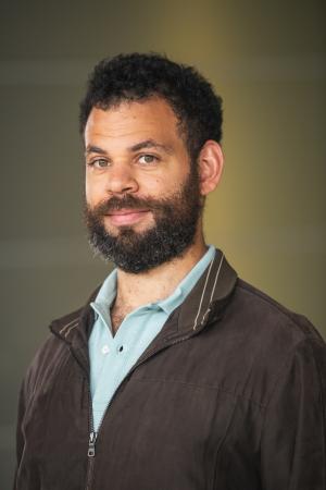A person, wearing a light blue collared shirt under a brown jacket. The background is out of focus and appears to be indoors.
