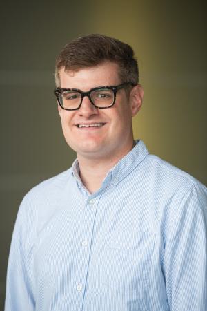 A person wearing a light blue, button-down shirt with vertical stripes. The background is a gradient from dark green to yellowish-green.