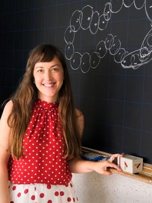 A person standing in front of a blackboard with a grid pattern and a drawing of a molecular structure made of circles and lines. The person is wearing a red polka dot top and skirt, holding a small box with designs. 
