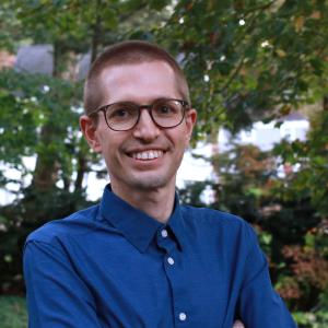 A person in a blue button-up shirt stands outdoors with trees and foliage in the background.