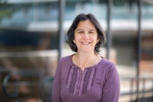 A person with short hair is photographed outdoors in front of a glass building. The person is wearing a long‑sleeved top with decorative vertical fabric patterns on the front. 