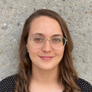 A person with shoulder‑length wavy hair stands in front of a textured concrete wall. The person is wearing a dark shirt with small white dots.