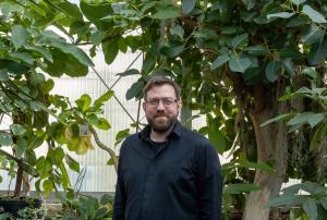 A person wearing a long‑sleeved dark shirt stands inside a greenhouse surrounded by large leafy plants and trees. The background includes translucent greenhouse panels.