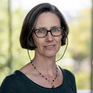 A person with short hair is shown from the shoulders up, wearing a dark top and a beaded necklace. The background is softly blurred greenery. 