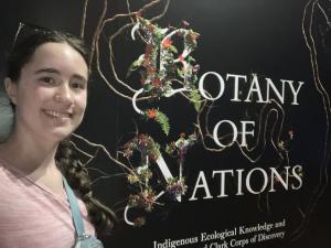 Person standing beside a “Botany of Nations” exhibit decorated with vines, branches, and flowers.