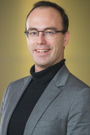 Professional headshot of a person wearing glasses, a gray blazer, and a black turtleneck against a soft background.