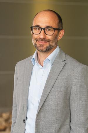 Professional headshot of a person wearing glasses, a light gray blazer, and a light blue shirt against a neutral background.