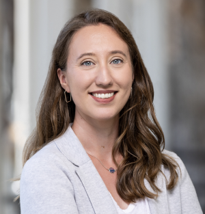 Professional headshot of a person with long dark hair wearing a cream blazer and white blouse against a neutral background.