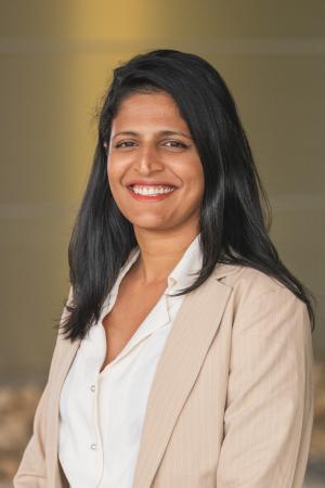 Professional headshot of a person with long brown hair wearing a light blazer, photographed against a softly blurred background.