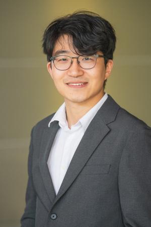Professional headshot of a person wearing glasses, a gray blazer, and a white shirt against a neutral background.