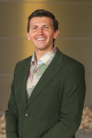 Professional headshot of a person in a dark green blazer and patterned shirt, photographed against a softly blurred background.