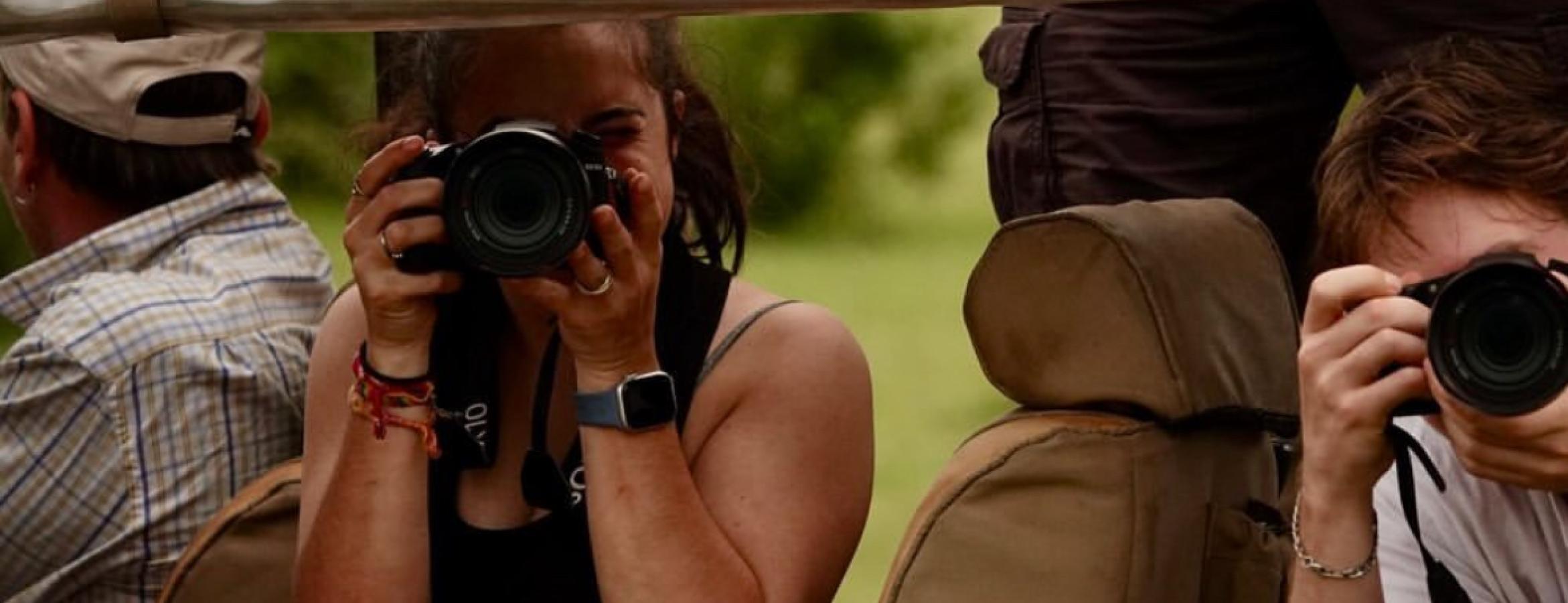 Three people seated in an open safari vehicle, two of them holding cameras and taking photographs, with green vegetation visible in the background.