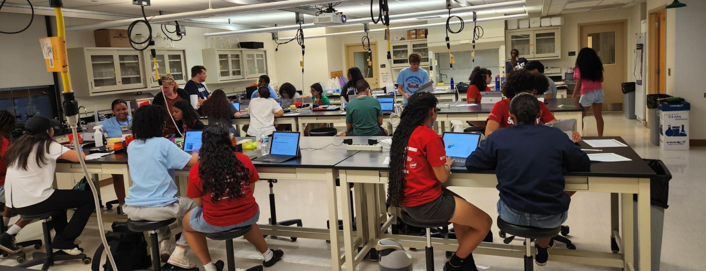 Students working at lab tables with laptops and papers in a bright science classroom.