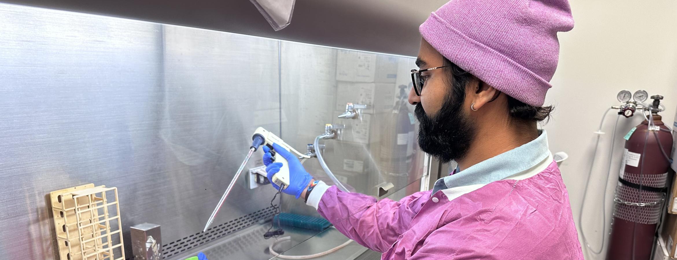 A person dressed in a bright pink protective lab gown and matching pink cap, working inside a laboratory biosafety cabinet. The individual is using a pipette to transfer liquid into a small container placed in an orange rack. The workspace includes lab equipment such as racks, boxes, and gas cylinders in the background.
