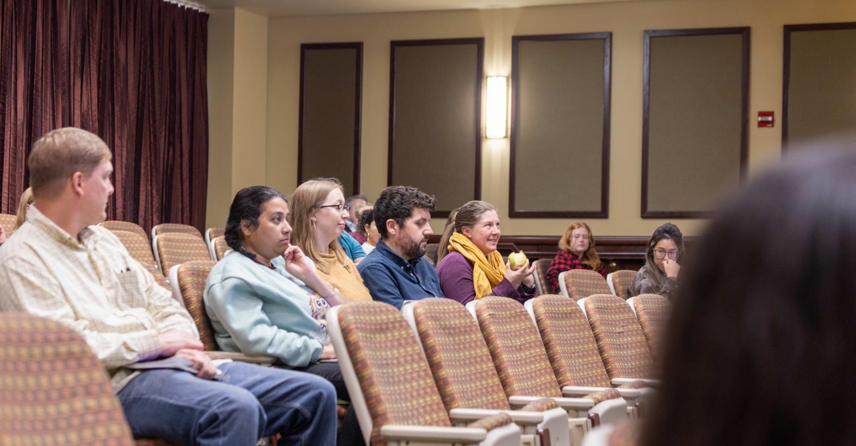 eople seated in rows of patterned chairs inside a lecture hall or auditorium.