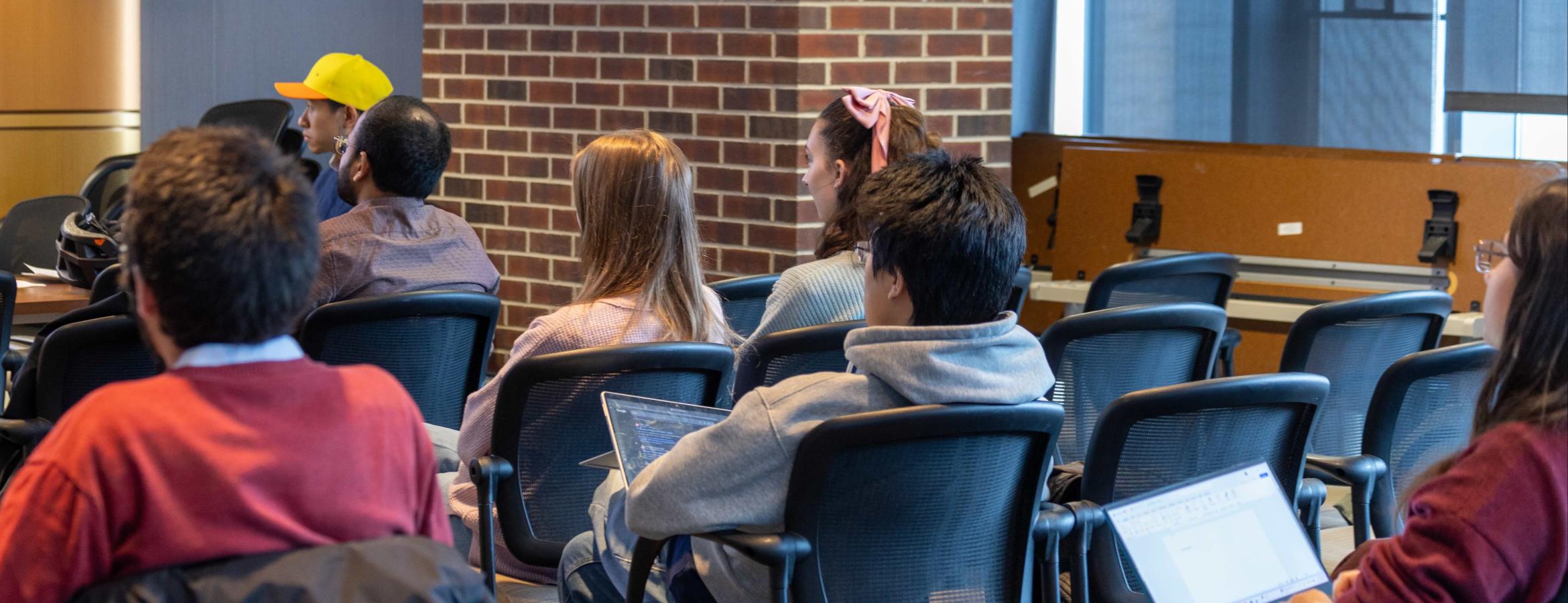 Group seated in chairs, some using laptops, in a classroom with brick walls and large windows.