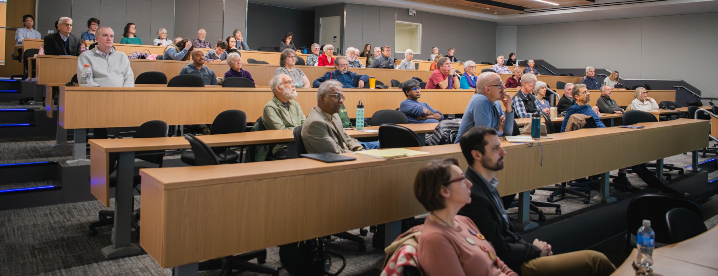 Audience seated in a modern lecture hall listening to a presentation, with tiered rows and ceiling‑mounted projectors.