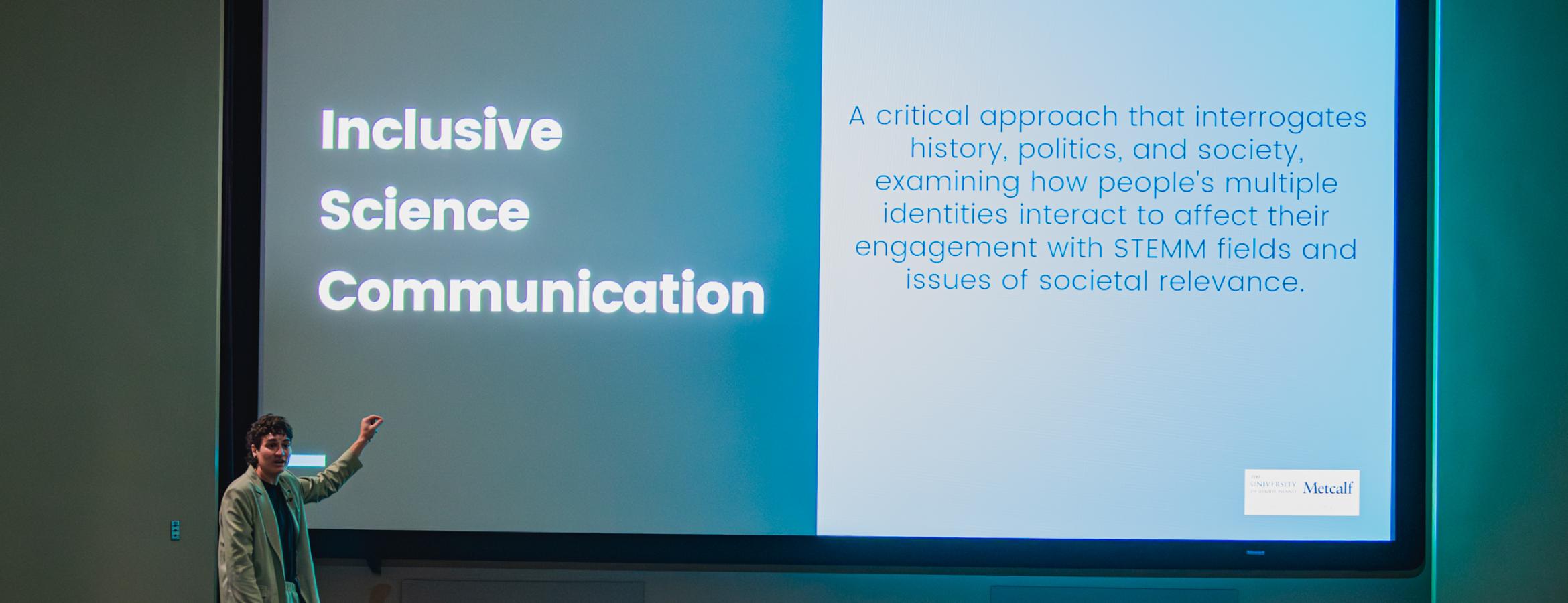 Presenter points to a slide titled “Inclusive Science Communication” while addressing an audience in a lecture hall.