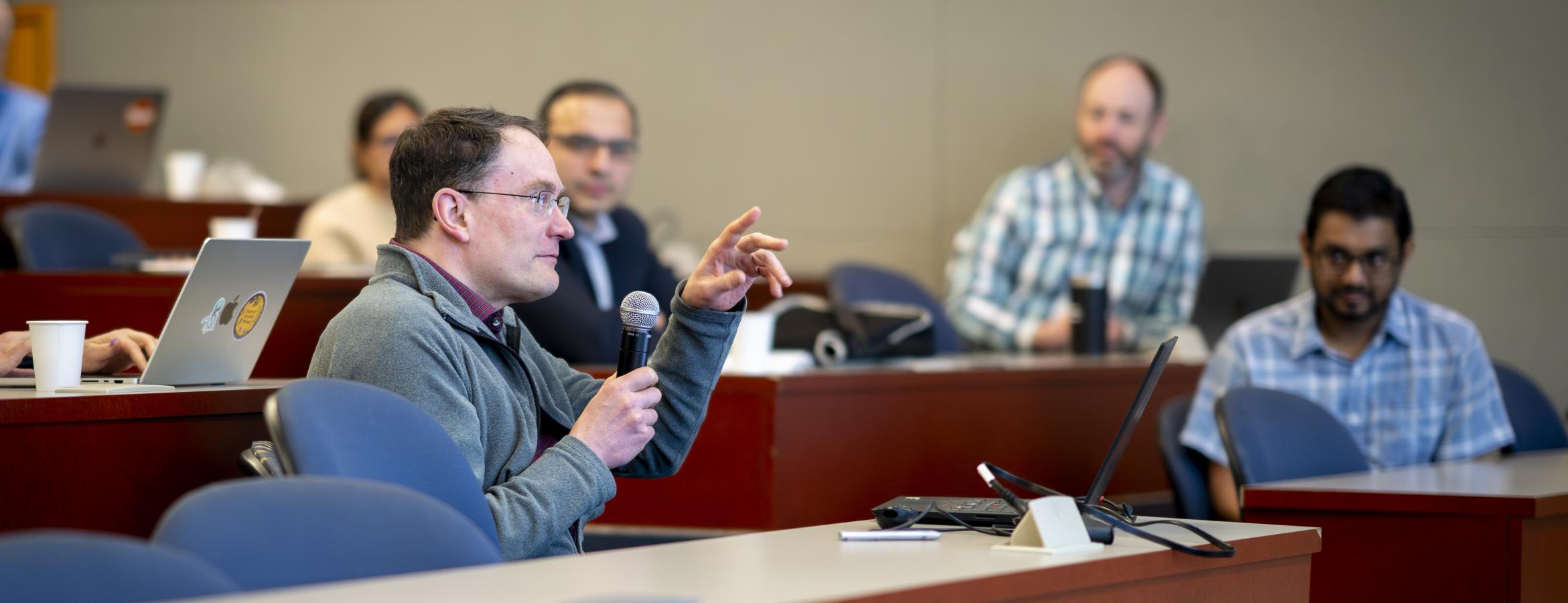 Audience member holds a microphone while speaking from tiered seating, other attendees and laptops visible in classroom.