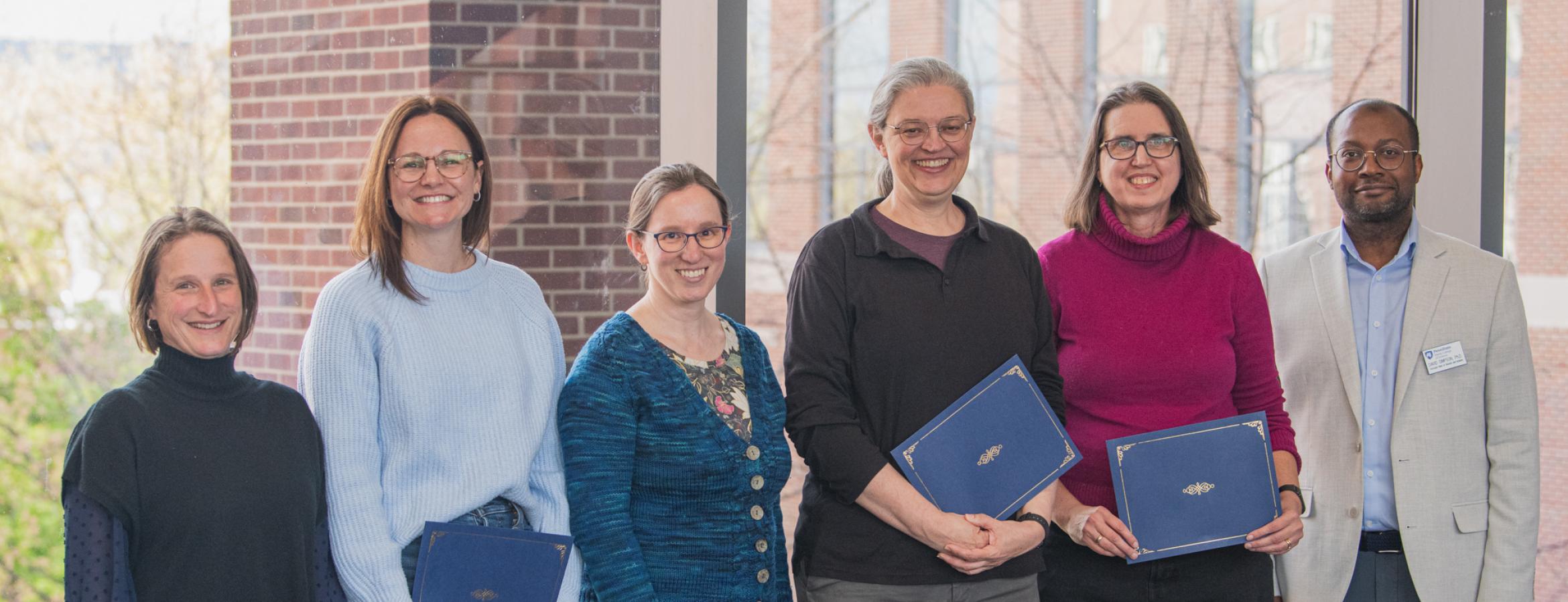 Six people stand indoors by large windows, several holding blue certificate folders, posed in a group for an awards photo.